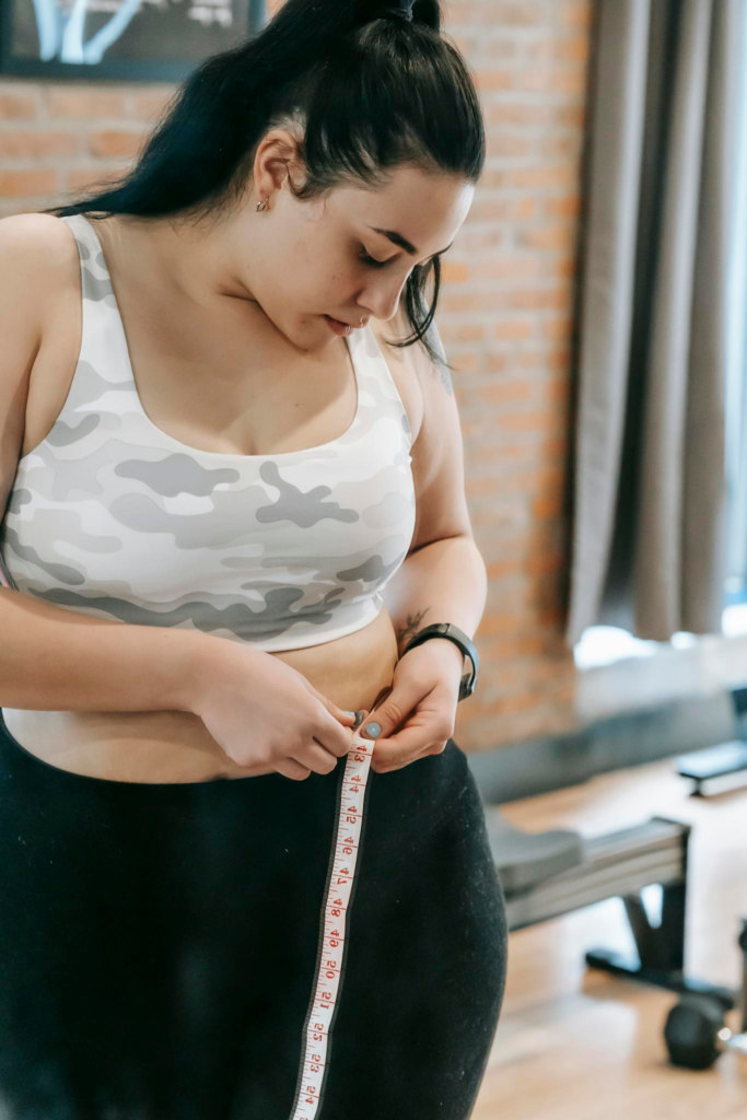 Young obese female using tape for evaluating size of belly while preparing for training in modern fitness center